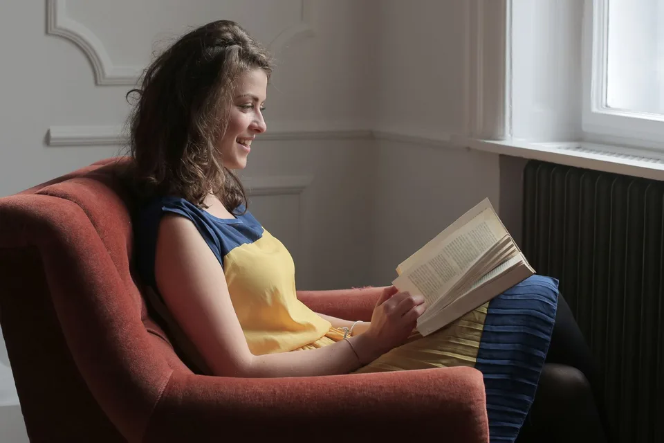 A person reading a paperback in an armchair beside a window