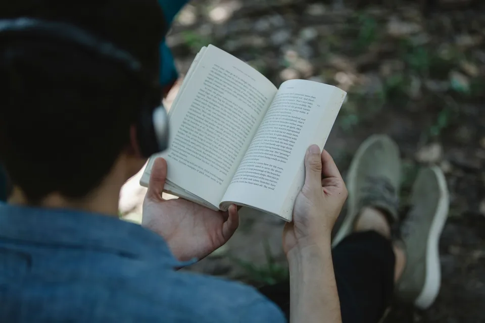 A person listening on headphones with an open book in a park